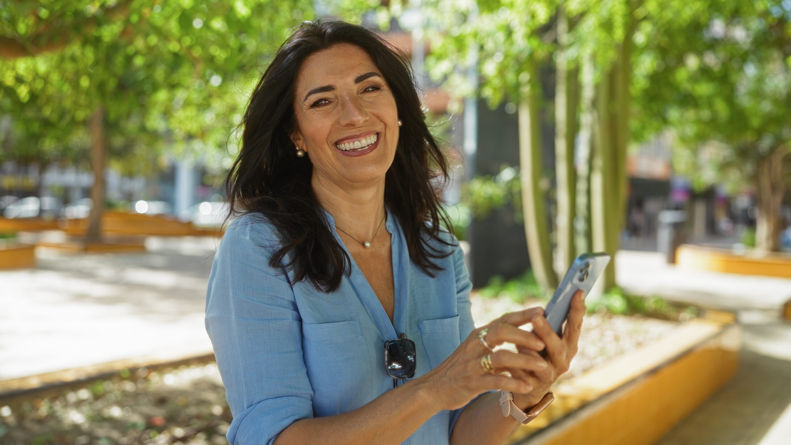 Hispanic,Woman,Smiling,And,Using,A,Mobile,Phone,Outdoors,In